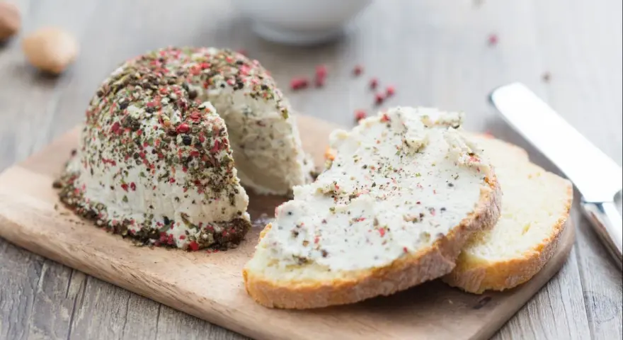 Queso vegano receta: un bloque de queso cremoso cubierto de pimienta y hierbas, servido con rebanadas de pan.