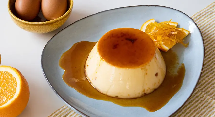 Delicioso flan de naranja con caramelo líquido, decorado con rodajas y ralladura de naranja. Huevos en un cuenco al fondo.