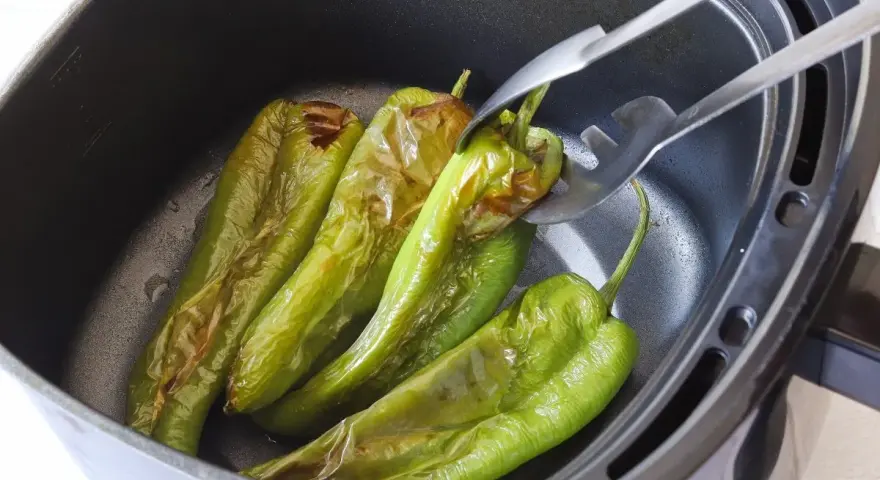 Pimientos verdes asándose en una freidora de aire. Unas pinzas metálicas sujetan uno de los pimientos, mostrando su piel ligeramente chamuscada.