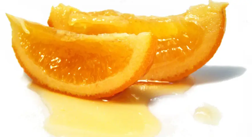 Dos rodajas de naranja confitada brillan con dulzura, bañadas en un almíbar dorado sobre fondo blanco.