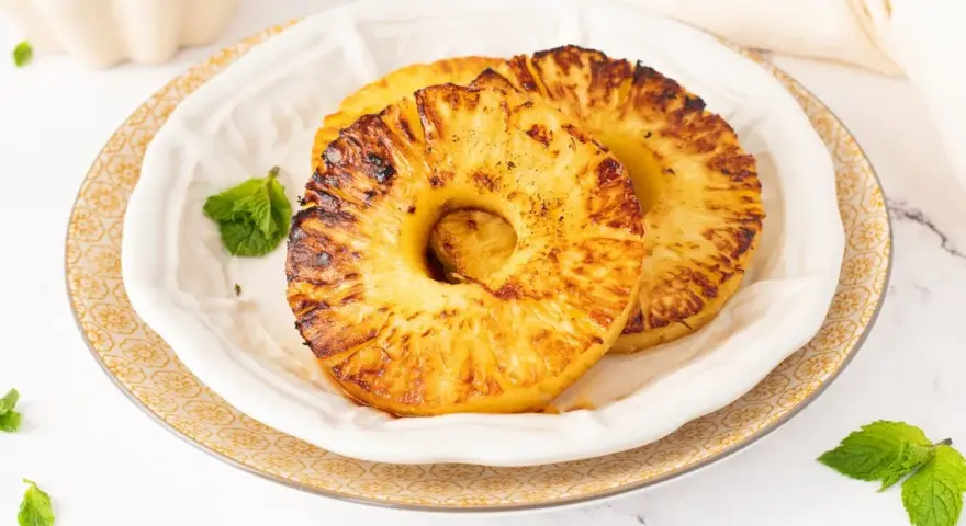 Dos rodajas de piña a la plancha, doradas y caramelizadas, servidas en un plato blanco con hojas de menta.