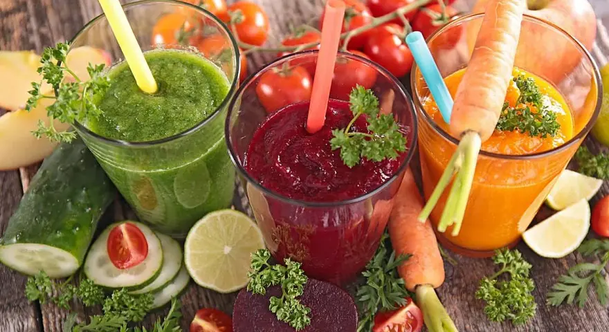 Tres zumos astringentes de colores vibrantes: verde, rojo y naranja, rodeados de ingredientes frescos como zanahorias, remolachas y tomates.