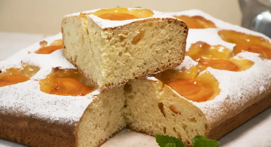 Deliciosa coca de albaricoque, cubierta de azúcar glas y decorada con trozos de fruta. Un trozo apilado revela su esponjosa miga.