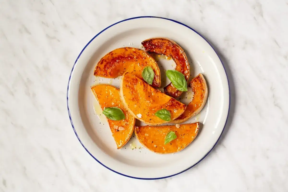 Rodajas de calabaza asada, glaseadas y con hojas de albahaca fresca, servidas en un plato blanco con borde azul sobre una superficie de m&aacute;rmol.