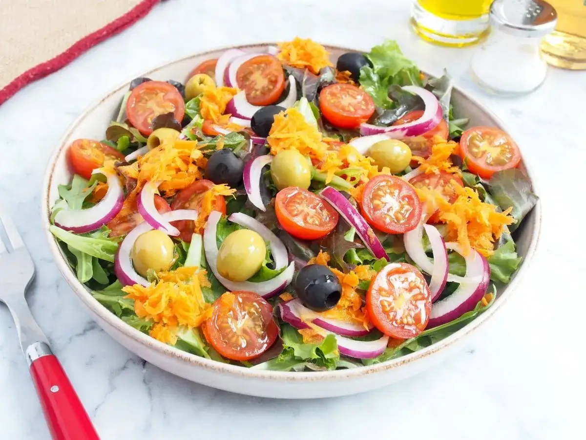Ensalada mediterr&aacute;nea vibrante con tomates cherry, aceitunas, cebolla morada y zanahoria rallada sobre hojas verdes frescas.