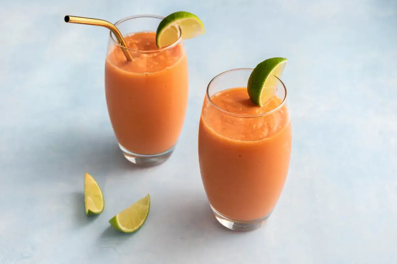 Dos vasos de zumo de papaya refrescante, adornados con rodajas de lima y pajitas doradas, listos para disfrutar.