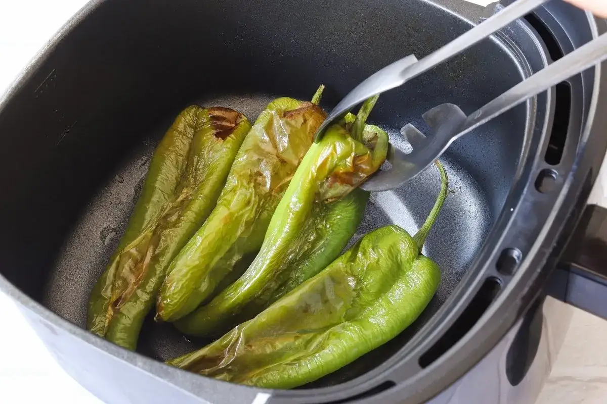 Pimientos verdes as&aacute;ndose en la freidora de aire. Unas pinzas voltean uno de los pimientos para que se cocine uniformemente.