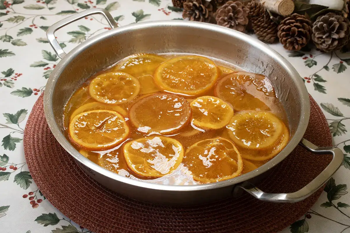 Rodajas de naranja confitada burbujeando en almíbar dentro de una sartén metálica, sobre un mantel festivo.
