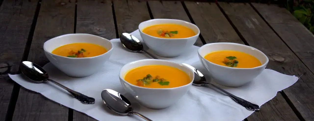 Cuatro tazones de sopa de calabaza cremosos, adornados con trocitos de verduras y perejil, esperan ser disfrutados sobre una servilleta blanca en una mesa de madera r&uacute;stica.