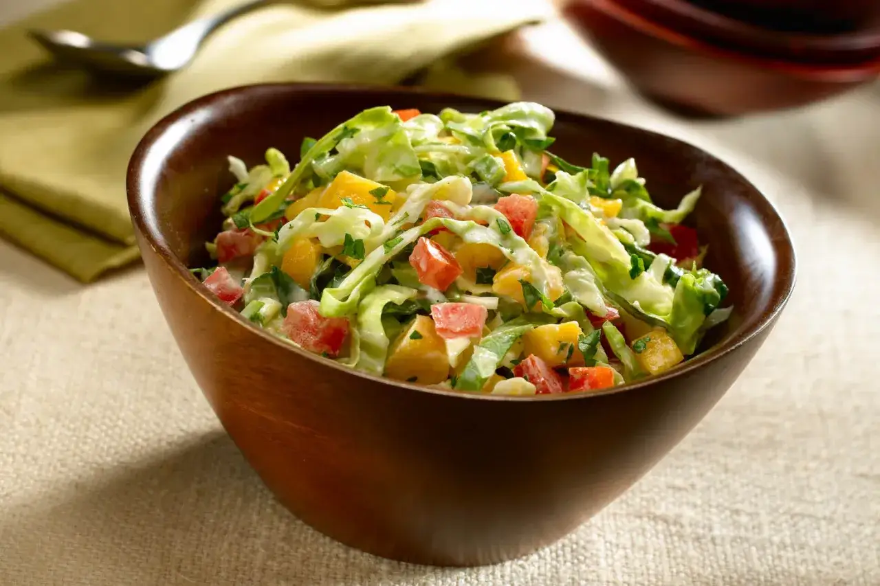 Ensalada tropical en un cuenco de madera, con trozos de mango, tomate y repollo, aderezada con una cremosa salsa.