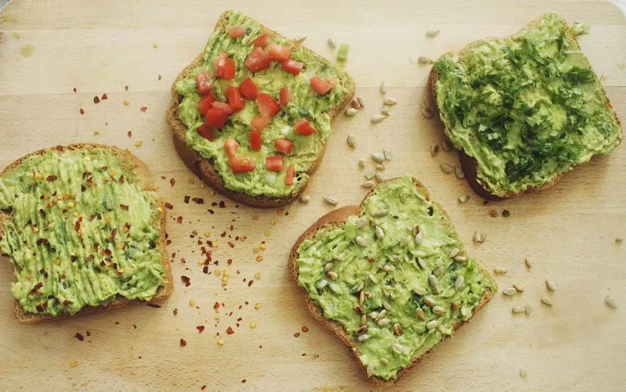 Cuatro tostadas de aguacate, una con tomate, otra con hierbas, y dos con semillas, esparcidas sobre una tabla de madera.