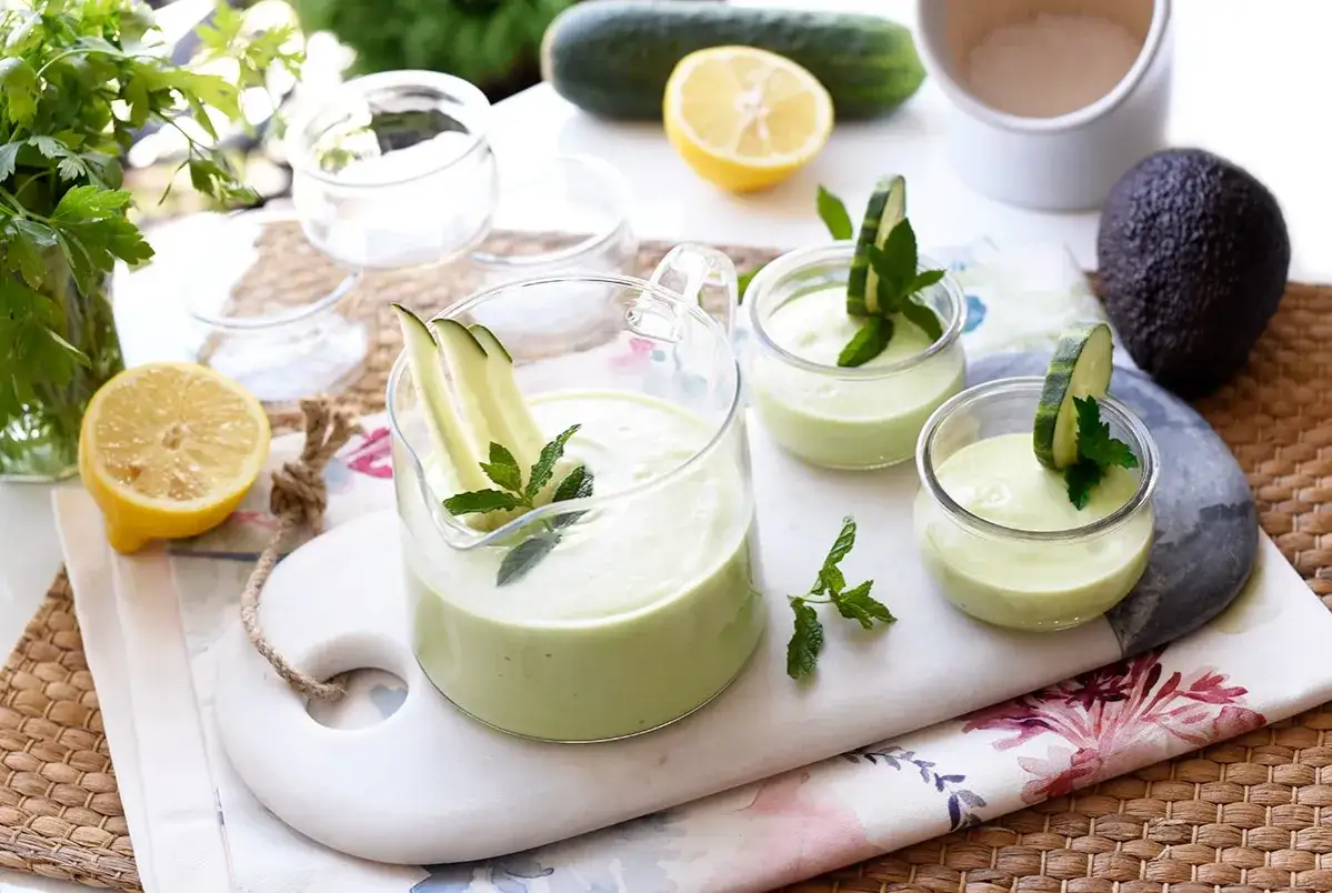 Refrescante gazpacho de aguacate servido en jarra y copas, decorado con pepino y menta. Ingredientes frescos como lim&oacute;n y perejil completan la escena.