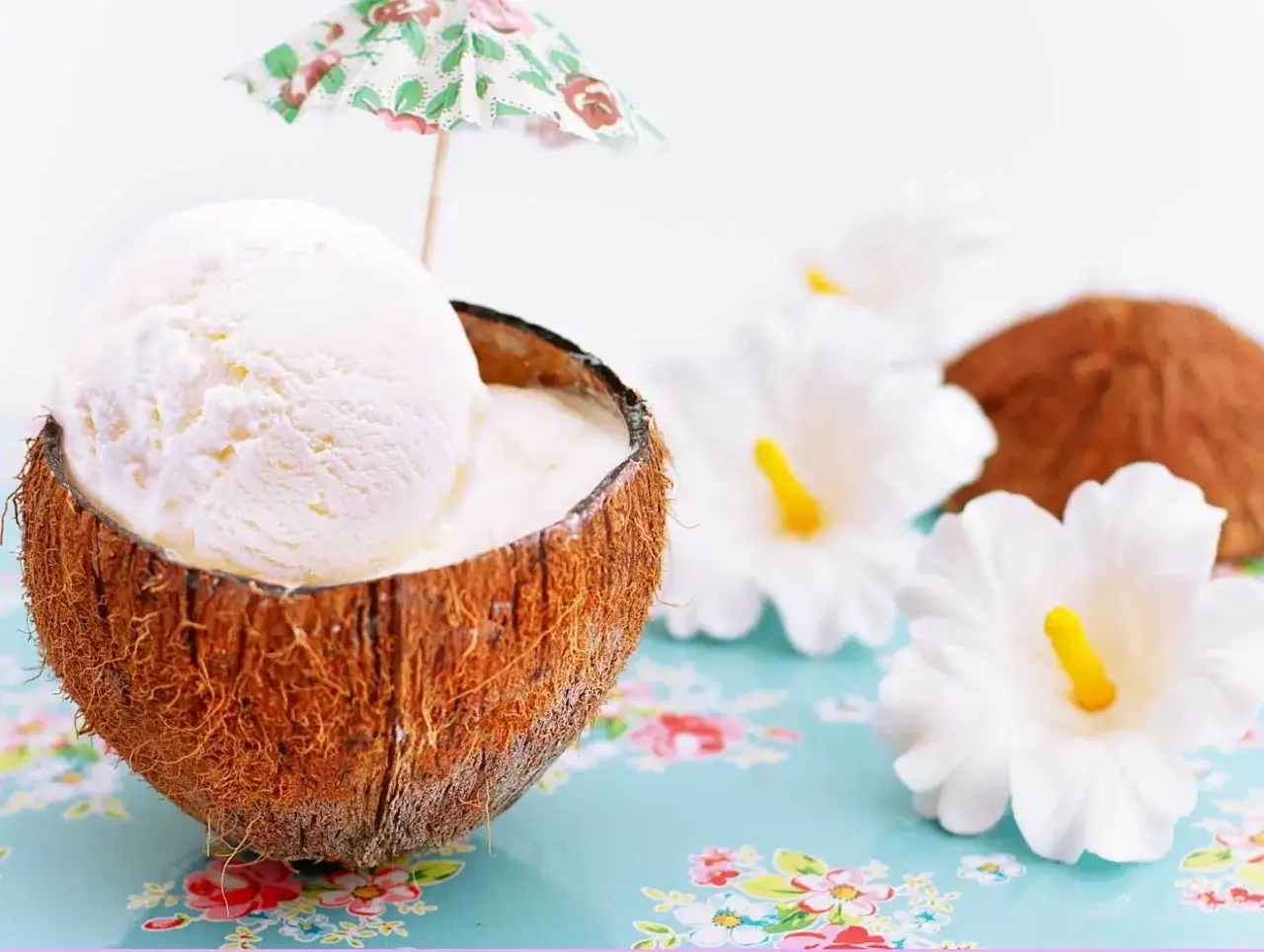 Delicioso helado con leche de coco servido en una c&aacute;scara de coco, decorado con una sombrilla tropical y flores blancas.