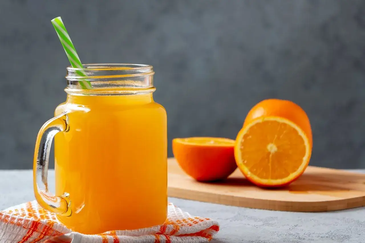 Jarra de jugo de naranja con pajilla verde y blanca, junto a naranjas frescas cortadas. Un refrescante ejemplo de para qu&eacute; sirve la naranja: &iexcl;vitamina C!