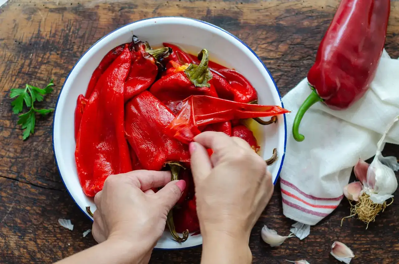Manos pelando pimientos asados en un cuenco blanco. Al lado, un pimiento rojo fresco, ajo y perejil.