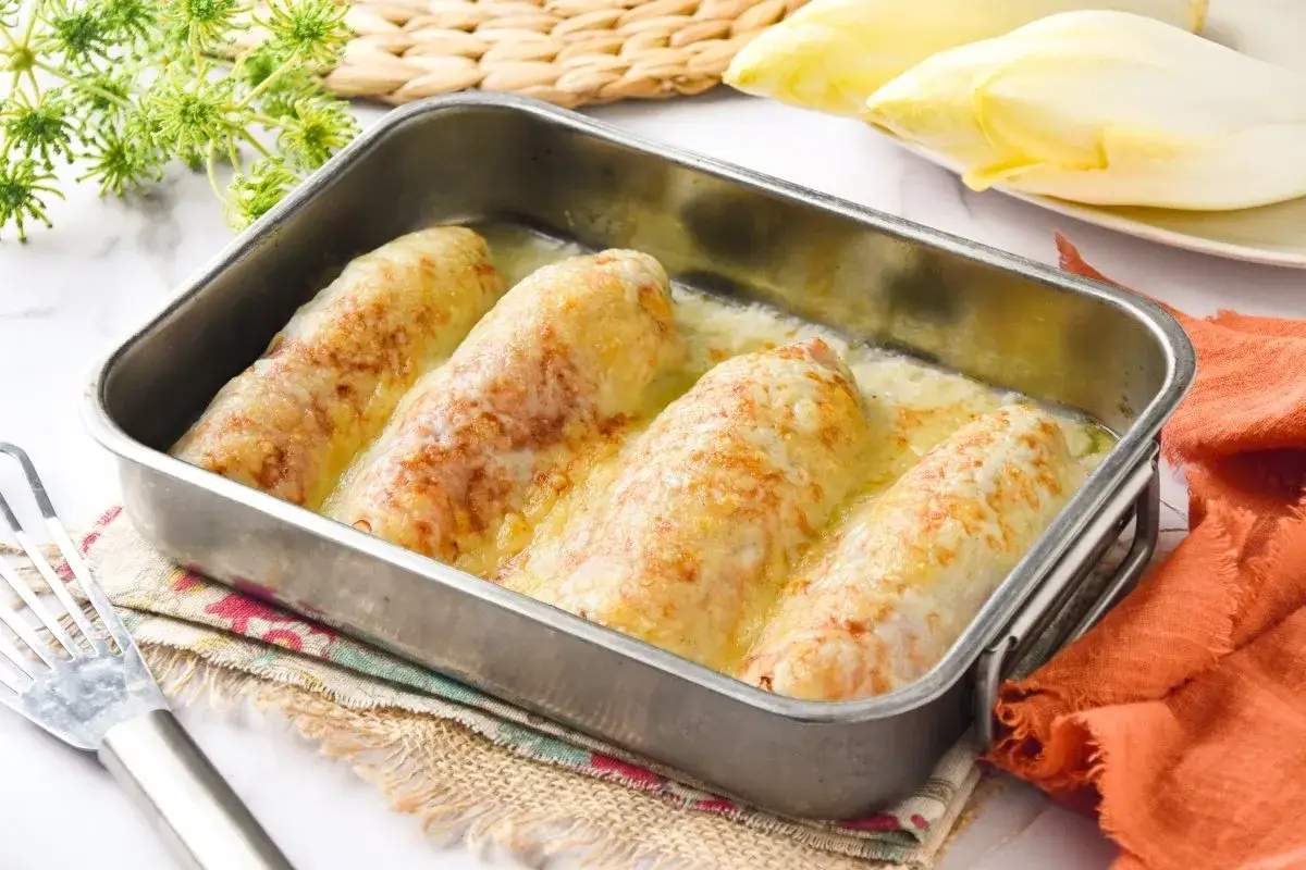 Cuatro rollos de endivias gratinadas con queso en una bandeja met&aacute;lica, listos para servir.
