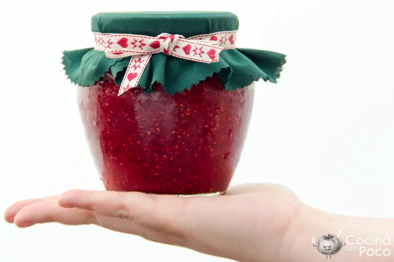 Mano sostiene un frasco casero de mermelada de frambuesa, decorado con tela verde y un lazo rojo con corazones.