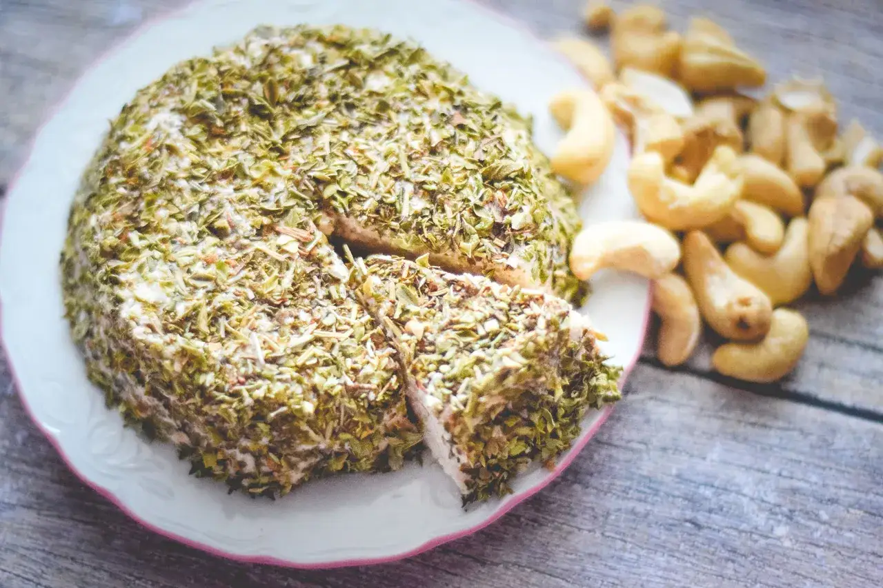 Queso vegano receta: un redondo y cremoso queso cubierto de hierbas, cortado en porciones, junto a anacardos.