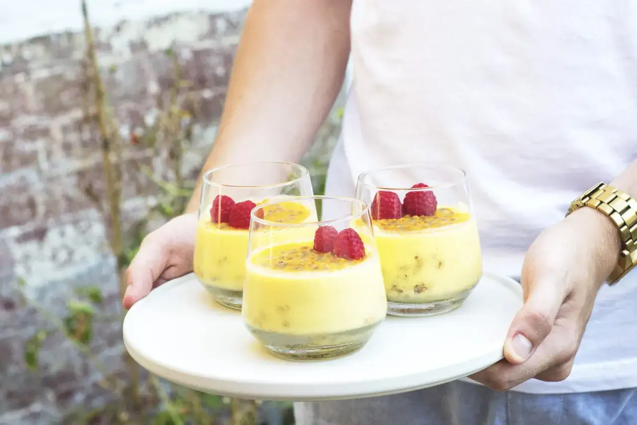Tres copas de cremoso mousse de maracuya, decoradas con frambuesas y semillas de maracuy&aacute;, servidas en una bandeja blanca.