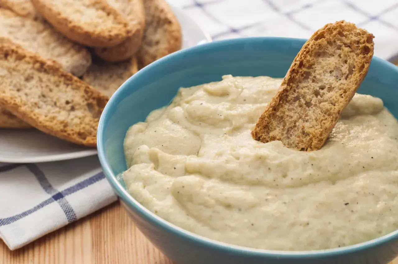 Un trozo de pan crujiente se sumerge en un cuenco azul con cremosa crema de berenjena.