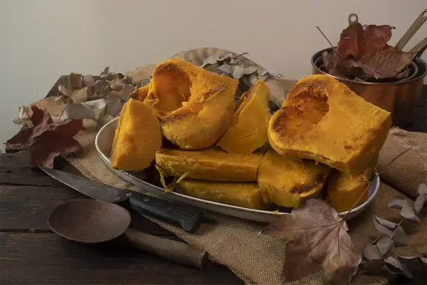 Trozos de calabaza asada, tierna y dorada, servidos en un plato blanco sobre arpillera. Acompañada de utensilios rústicos y hojas secas, evoca el sabor del otoño.