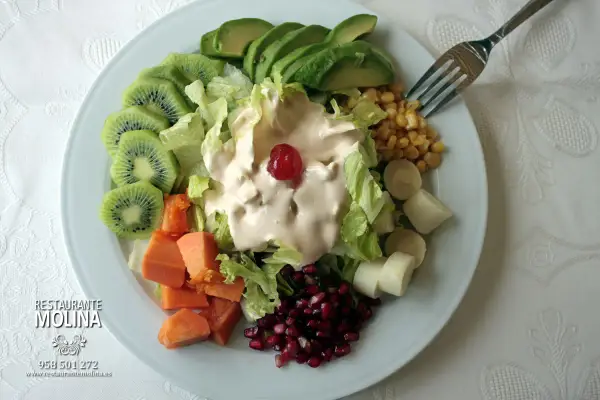 Ensalada tropical con aguacate, kiwi, papaya, palmitos, maíz y granada, cubierta de aderezo cremoso y una cereza.