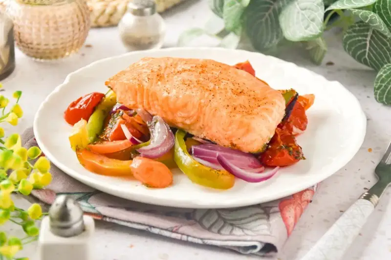 Un jugoso salmón al horno reposa sobre una cama de verduras asadas: tomates, pimientos y cebolla morada.